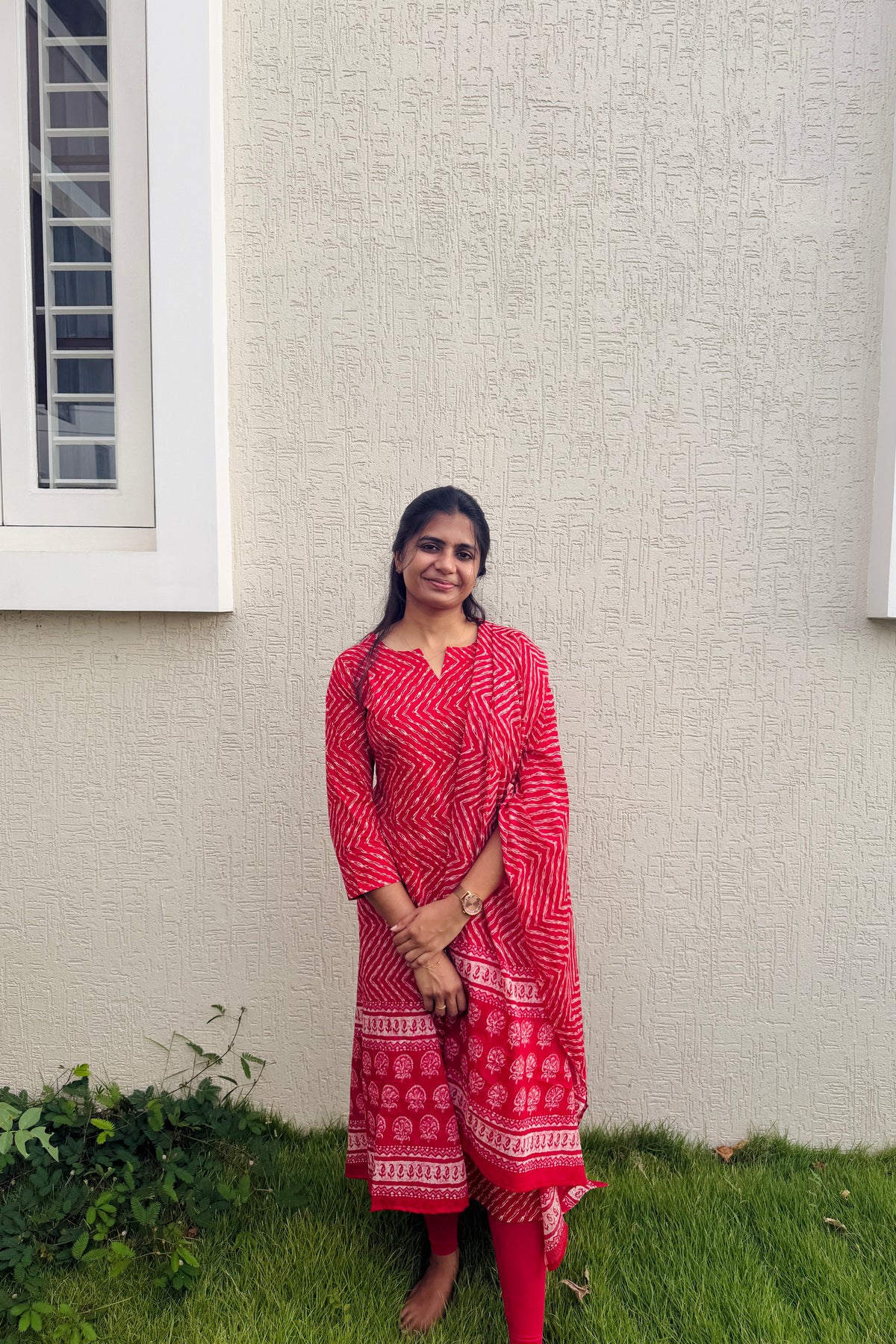 A woman stands outdoors in front of a textured beige wall and green grass, wearing a red and white patterned kurta with matching red trousers.