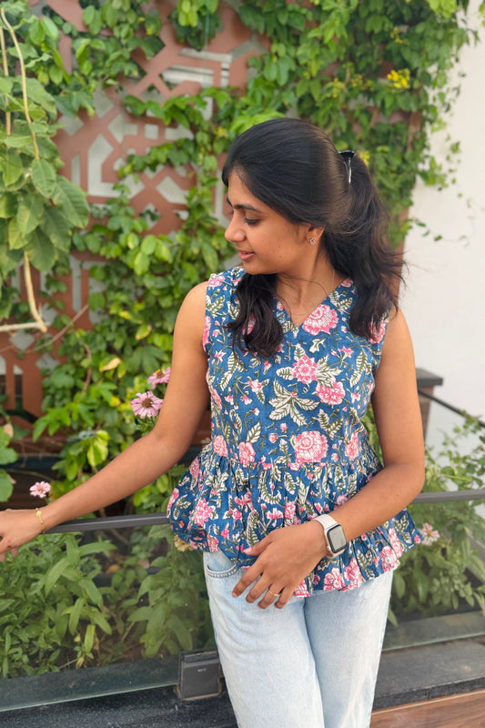 A woman wearing a sleeveless navy blue floral peplum top and light wash jeans, standing outdoors in front of a green leafy wall.