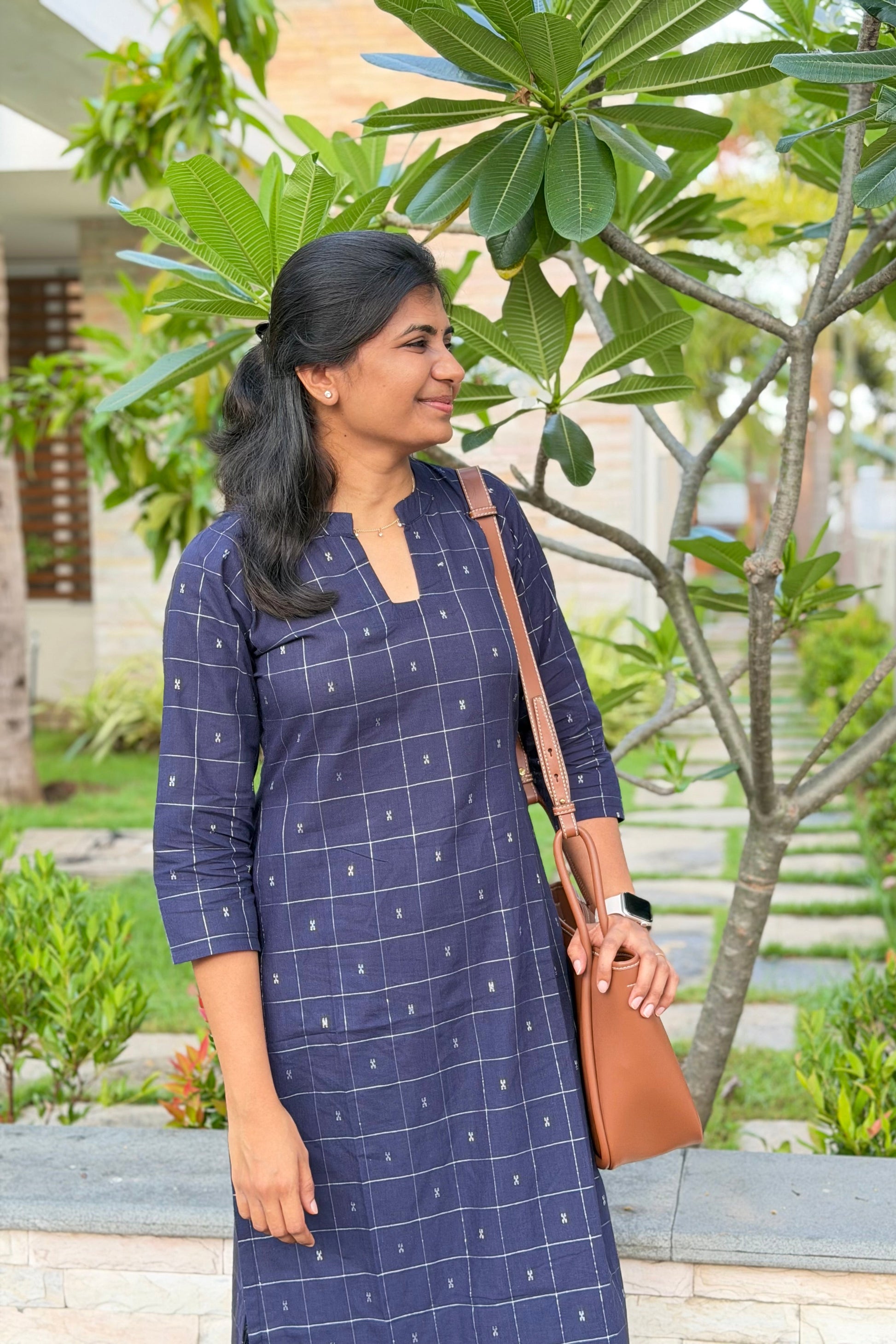 Woman in a blue checkered dress holding a brown bag outdoors.