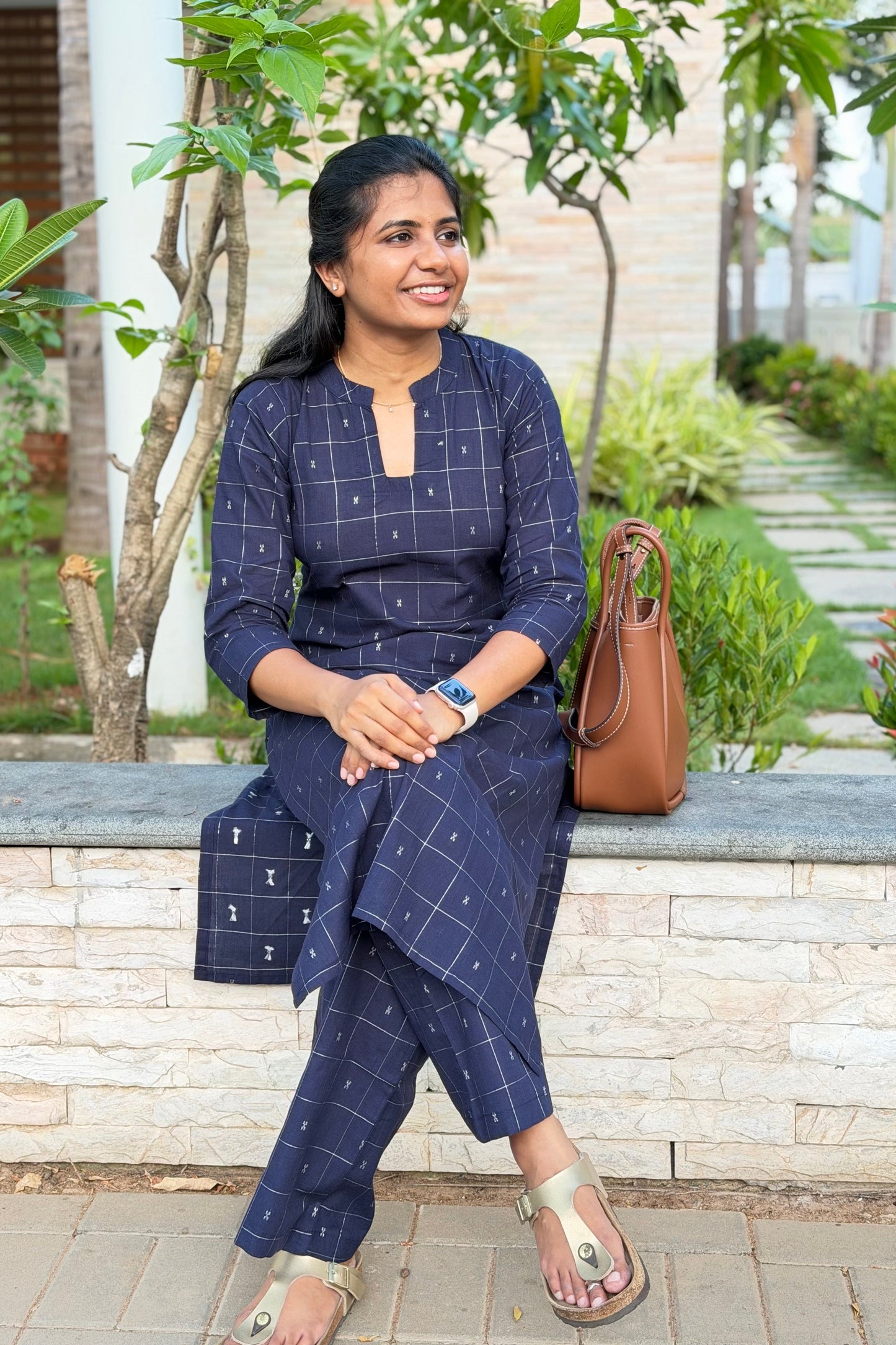 Woman in a blue checkered outfit sitting outdoors with a brown handbag.